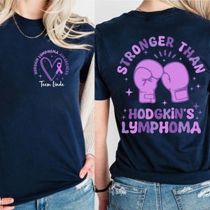 May include: Navy blue t-shirt with Hodgkin's Lymphoma awareness design. The front features a purple heart and ribbon with text. The back displays boxing gloves and the words "STRONGER THAN" and "HODGKIN'S LYMPHOMA" in purple.