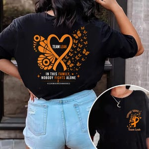 Könnte beinhalten: Ein schwarzes T-Shirt mit einem großen orangefarbenen Schmetterlingsflügel-Design auf der Rückseite, das ein herzförmiges Band mit dem Text TEAM LEAH enthält. Darunter steht IN THIS FAMILY, NOBODY FIGHTS ALONE und #LEUKEMIACANCER AWARENESS.