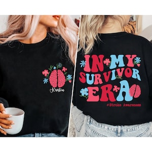 May include: Black sweatshirt with a pink brain graphic and the text "In My Survivor Era" in pink, blue, and red. The text "#Stroke Awareness" is printed below the graphic. The sweatshirt has a floral design with a pink brain graphic and the name "Kristen" printed below the graphic.