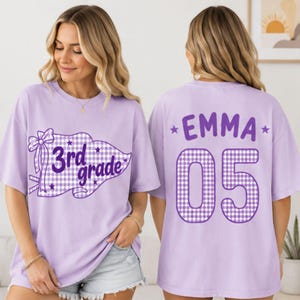 Könnte beinhalten: Ein hellviolettes T-Shirt mit einer violett-weiß karierten Wimpelgrafik, einer Schleife und dem Text 3rd grade auf der Vorderseite sowie dem Namen EMMA und der Zahl 05 im violett-weißen Karomuster auf der Rückseite des Shirts.