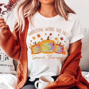 Può includere: T-shirt bianca con la scritta "Harvesting Words This Fall" e "Speech Therapy". La maglietta presenta tre zucche arancioni con grafici colorati di terapia del linguaggio. Foglie autunnali e ghiande sono sparse intorno alle zucche.