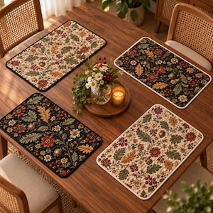 May include: Four rectangular placemats featuring a quilted texture and a botanical pattern of oak leaves, acorns, and flowers in shades of red, green, and gold. Two placemats have a cream background, and two have a dark black background.