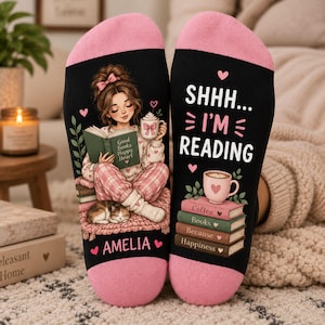 May include: A pair of black socks with pink cuffs. The left sock features an illustration of a person reading a book with a cat, and the name AMELIA. The right sock displays the text SHHH... I'M READING above a stack of books and a coffee mug.