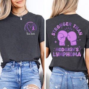 May include: Dark gray t-shirt with Hodgkin's Lymphoma awareness design. Front features a purple heart and ribbon with text. Back displays purple boxing gloves and the words "Stronger Than Hodgkin's Lymphoma."