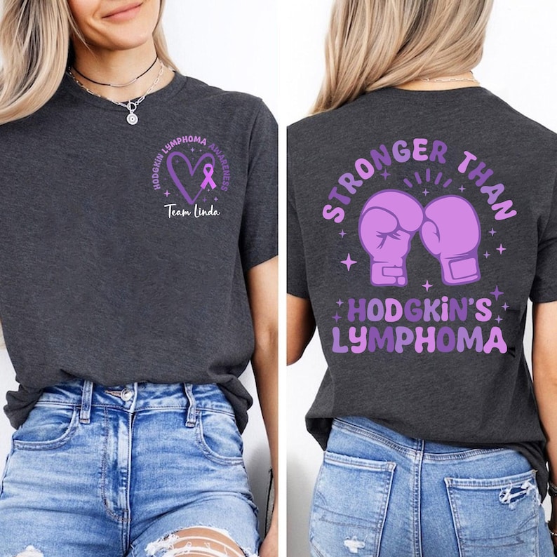 May include: Dark gray t-shirt with Hodgkin's Lymphoma awareness design. Front features a purple heart and ribbon with text. Back displays purple boxing gloves and the words "Stronger Than Hodgkin's Lymphoma."