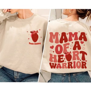 Pode incluir: Uma camisa de moletom bege com um coração vermelho e o texto "Team Ashley" na frente. A parte de trás da camisa de moletom tem o texto "Mama of a Heart Warrior" em letras vermelhas e rosa com um coração vermelho.