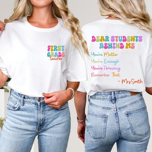 May include: Two white t-shirts with text printed on them. The first shirt says "First Grade Teacher" in rainbow colors. The second shirt says "Dear Students Behind Me You're Matter You're Enough You're Amazing Remember That - Mrs. Smith" in rainbow colors.