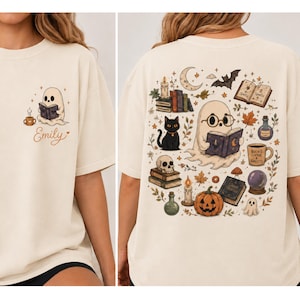Puede incluir: Una camiseta color hueso con un peque&ntilde;o gr&aacute;fico en el frente de un fantasma leyendo un libro con el nombre Emily y una taza de caf&eacute;. La parte posterior muestra una gran ilustraci&oacute;n de un fantasma con gafas leyendo, rodeado de libros, un gato, una calabaza y velas.