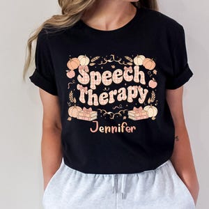Peut inclure: T-shirt noir avec un motif sur le thème de l'automne. Le motif comprend les mots "Speech Therapy" dans une police à carreaux, entourés de citrouilles, de feuilles et du nom "Jennifer".