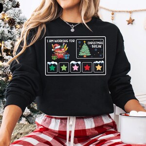 May include: Black sweatshirt featuring a graphic of a progress bar for Christmas break. The bar is nearly full, with a red star showing the current progress. The text reads "I am working for Christmas Break".