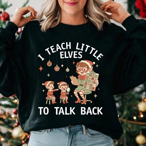 May include: Black sweatshirt with the text "I TEACH LITTLE ELVES TO TALK BACK" and a graphic of elves. The graphic shows an elf teacher with glasses and a clipboard, teaching two elves. Christmas ornaments and stars decorate the design.