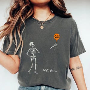 Peut inclure: Un t-shirt gris anthracite à manches courtes avec un motif de squelette blanc, un bras squelettique tenant un ballon orange en forme de citrouille d'Halloween et le texte "Well, shit..." imprimé en blanc sous le motif.