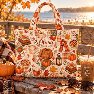 May include: A quilted tote bag featuring a seasonal autumn print with pumpkins, fallen leaves, books, a jumper, a scarf, a lantern, and mugs. The name Olivia is printed in the centre. The bag has two top handles and a rectangular structure.