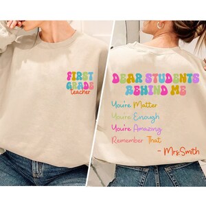 May include: A beige sweatshirt with the text "FIRST GRADE teacher" on the front and "DEAR STUDENTS BEHIND ME You're Matter You're Enough You're Amazing Remember That - Mrs. Smith" on the back.