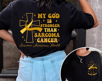 May include: Black t-shirt with a gold cross and ribbon design. The text reads "MY GOD IS STRONGER THAN SARCOMA CANCER" and "Sarcoma Awareness Month." A smaller image shows a similar design with "TEAM EMILY."