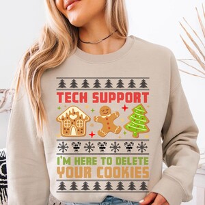 Op de afbeelding: Beige sweatshirt met een feestelijk ontwerp. Het ontwerp bevat een peperkoekhuisje, een peperkoekmannetje en een kerstboom. De tekst "TECH SUPPORT" en "I'M HERE TO DELETE YOUR COOKIES" worden in rood en groen weergegeven.