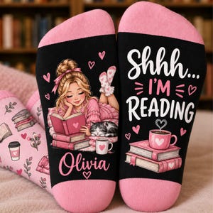 May include: A pair of black and pink socks featuring a graphic of a person with blonde hair reading a book next to a cat. The text reads Shhh... I'M READING and Olivia. The design includes pink hearts, stacks of books, and a pink coffee mug.