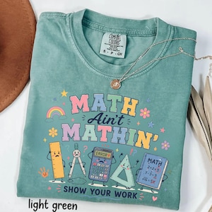 May include: A light green t-shirt featuring a graphic with the text "MATH AIN'T MATHIN' SHOW YOUR WORK" and cartoon illustrations of a ruler, compass, calculator, triangle, and notebook. The shirt has a crew neck and a Comfort Colors brand label.