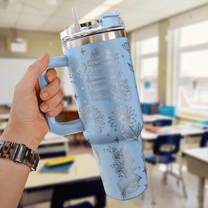 May include: A light blue insulated tumbler with a handle, straw, and lid. The tumbler is decorated with floral and book illustrations in silver. The tumbler is held in a hand, and the background is a classroom.