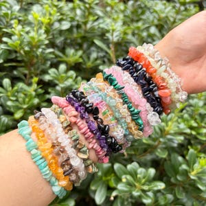 May include: A stack of multiple stretch bracelets made from various polished gemstone chips in colors including turquoise, orange, clear, pink, purple, black, green, and red, worn together on a wrist.