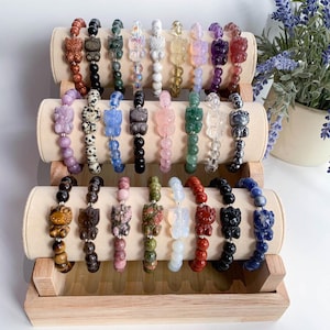 May include: A display of 18 colorful gemstone bracelets with cat charms. Each bracelet is made with different types of stones, including amethyst, rose quartz, tiger's eye, and lapis lazuli. The bracelets are displayed on a wooden stand.