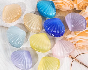 May include: A collection of colorful, translucent seashell-shaped decorative items. The shells are in various pastel colors, including blue, yellow, pink, and purple. The shells are arranged on a white surface with floral accents in the background.