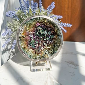 Puede incluir: Un espécimen de cristal redondo, iridiscente y multicolor sobre un soporte plateado. El cristal está compuesto por pequeños cristales de forma cuadrada que brillan con un arcoíris de colores.