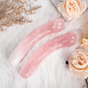Puede incluir: Dos herramientas de masaje de cuarzo rosa con un diseño curvo. Las herramientas son de color rosa suave y descansan sobre una superficie blanca y esponjosa. Tienen un acabado liso y pulido, diseñadas para masajes faciales.