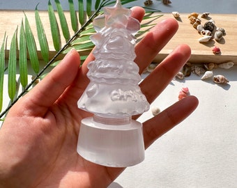 May include: A translucent Christmas tree figurine with a star on top, held in a hand. The tree has a tiered design with heart-shaped cutouts and the word "Christmas" etched on it. The base is a rectangular shape.