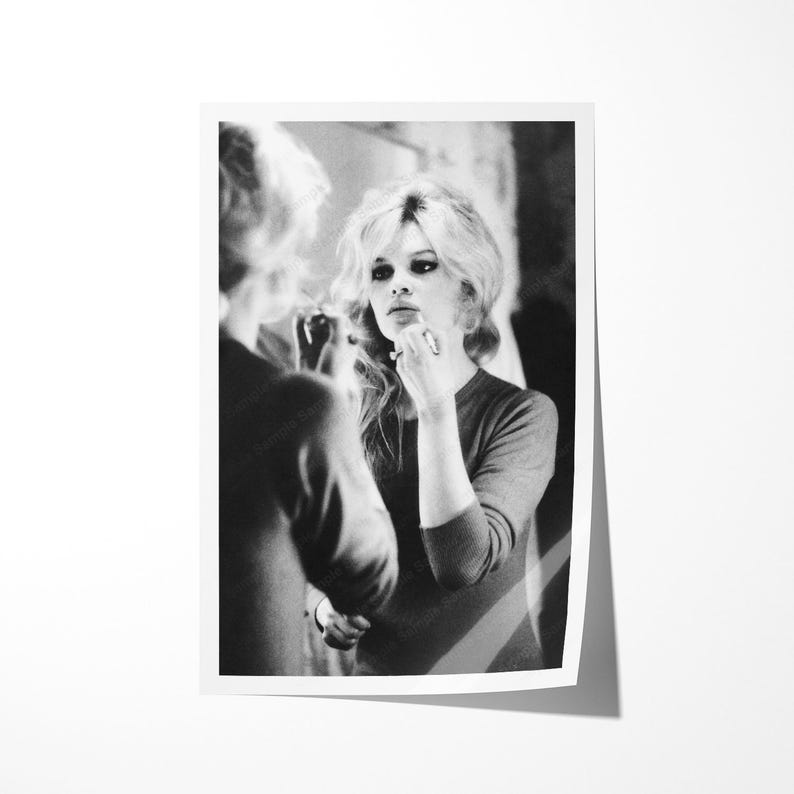 May include: Black and white photograph of a woman applying makeup in front of a mirror. The woman has blonde hair and is wearing a long-sleeved top. The image is a portrait, with the woman's reflection visible.