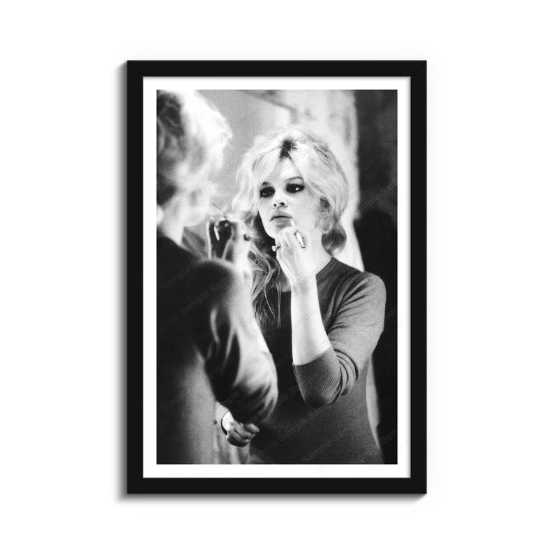 May include: Black and white framed print of a woman applying makeup in front of a mirror. The woman has blonde hair and is wearing a long-sleeved top. The image is a vintage style photograph.