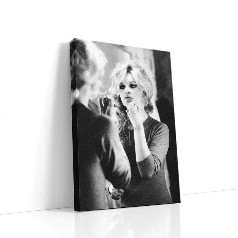 May include: Black and white canvas print of a woman applying makeup in front of a mirror. The woman has blonde hair and is wearing a long-sleeved top. The image is a portrait and is leaning against a white wall.