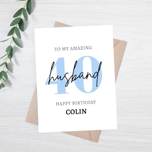 May include: A white birthday card with the text "TO MY AMAZING husband 40 HAPPY BIRTHDAY COLIN" in black and light blue. The card is on a brown envelope, with green leaves in the background.