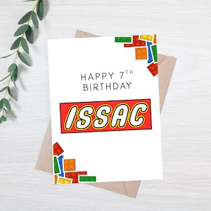 May include: A white birthday card with the text "HAPPY 7TH BIRTHDAY" and the name "ISSAC" in a red and yellow block design. Colorful toy blocks are in the corners. The card is on a brown envelope.