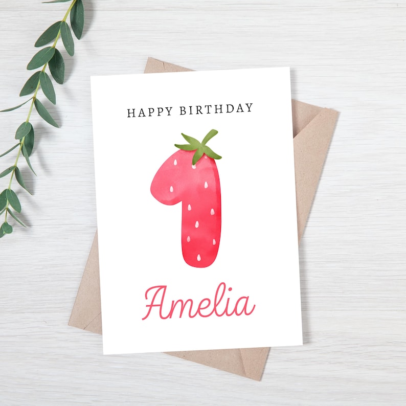 May include: A white card with the text "Happy Birthday" and the number "1" in the shape of a red strawberry with green leaves. The name "Amelia" is written in pink below the strawberry.