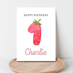 May include: A white card with the words "Happy Birthday" printed in black at the top. A red strawberry shaped like the number one is in the center of the card. The name "Charlie" is printed in cursive below the strawberry.
