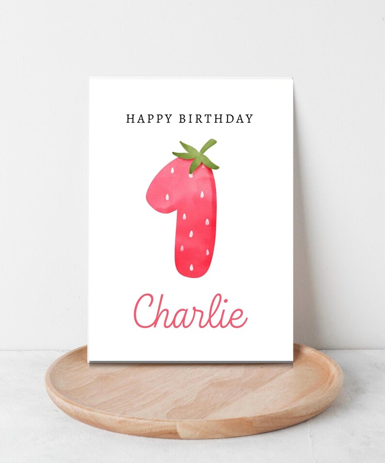 May include: A white card with the words "Happy Birthday" printed in black at the top. A red strawberry shaped like the number one is in the center of the card. The name "Charlie" is printed in cursive below the strawberry.