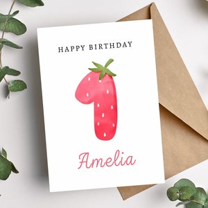May include: A white card with the text "Happy Birthday" and the number "1" in the shape of a red strawberry with green leaves. The name "Amelia" is written in pink below the strawberry.