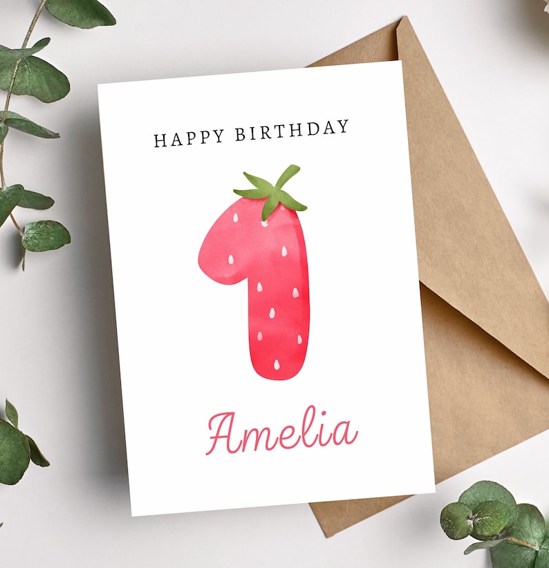 May include: A white card with the text "Happy Birthday" and the number "1" in the shape of a red strawberry with green leaves. The name "Amelia" is written in pink below the strawberry.