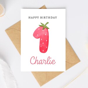 May include: A white card with the text "Happy Birthday" and a red watercolor strawberry shaped like the number one. The name "Charlie" is written in cursive below the number.
