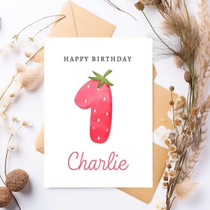 May include: A white card with the text "Happy Birthday" and a red strawberry shaped like the number one. The name "Charlie" is written below the number one.