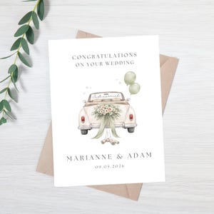May include: A wedding card with a vintage car illustration. The car is decorated with flowers and a ribbon. The card reads "Congratulations on your wedding" and includes the names "Marianne & Adam" and the date "09.05.2026".