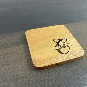 May include: A wooden coaster with a personalized design. The coaster features a cursive letter "C" with a line through the middle and the name "Camila" written below the line.