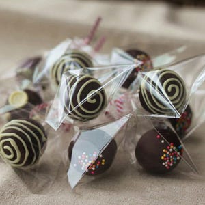 May include: Close-up of chocolate cake pops in clear cellophane bags. Some pops are decorated with white swirls, others with colorful sprinkles. The treats are arranged on a neutral-colored surface.