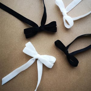 May include: Four velvet ribbon bows in black and white colours. Two bows are solid black and two are solid white. Each bow features a central knot with two long, trailing ribbon tails extending from the centre, creating a classic bow shape.