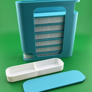 Puede incluir: Un pastillero semanal de plástico turquesa con siete compartimentos etiquetados para cada día de la semana. Se ve una bandeja blanca para pastillas para el domingo, etiquetada como "MAÑANA" y "NOCHE". También hay una tapa turquesa.