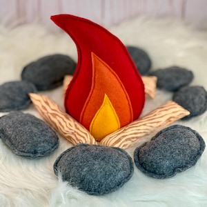 May include: A plush campfire set featuring a central flame made of layered red, orange, and yellow felt. The flame is surrounded by several soft, grey felt stones and light-coloured fabric logs printed with a brown wood grain pattern.