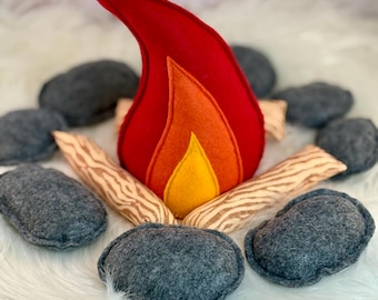 May include: A plush campfire set featuring a central flame made of layered red, orange, and yellow felt. The flame is surrounded by several soft, grey felt stones and light-colored fabric logs printed with a brown wood grain pattern.