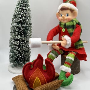 Könnte beinhalten: Eine Weihnachts-Elfenpuppe in einem rot-grünen Outfit sitzt auf einem Baumstamm und brät einen Marshmallow über einem Filz-Lagerfeuer. Der Elf hält einen Holzstab mit einem Marshmallow am Ende. Der Elf trägt einen grün-weiß gestreiften Schal und eine rot-grüne Mütze. Im Hintergrund steht ein kleiner künstlicher, schneebedeckter Tannenbaum.