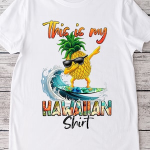 May include: White t-shirt with a graphic of a pineapple wearing sunglasses and doing the dab while surfing. The text "This is my Hawaiian Shirt" is printed below the pineapple.